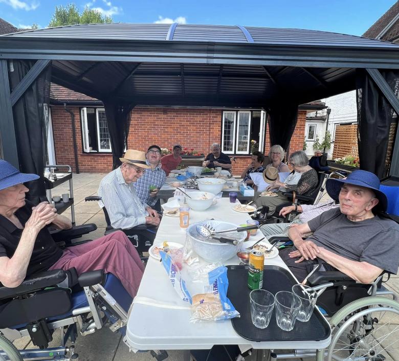 nursing home residents sat around tables with food on top outside in a courtyard under a marquee