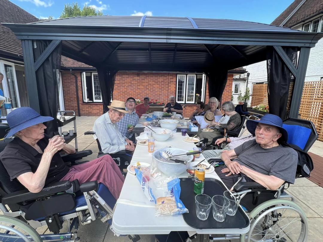 nursing home residents sat around tables with food on top outside in a courtyard under a marquee