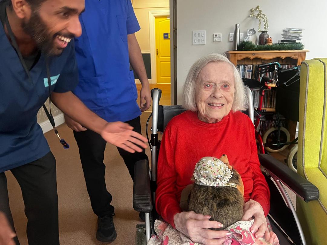 Smiling elderly woman in a red sweater holding a soft toy, seated in a wheelchair, surrounded by care staff