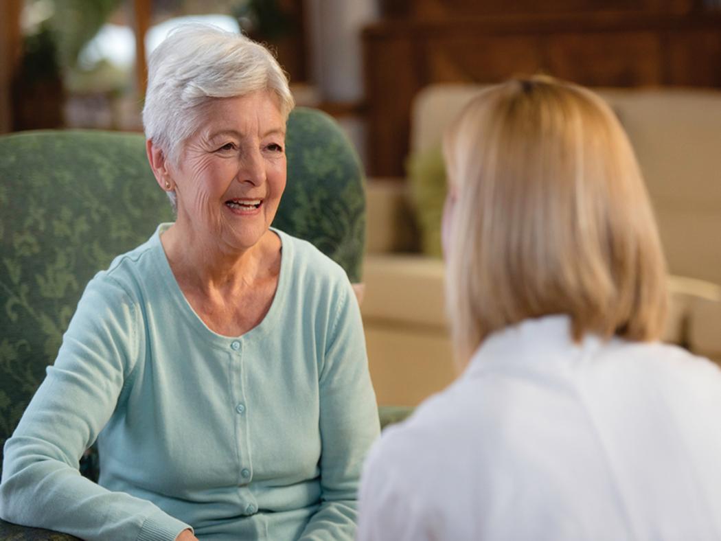 elderly lady smiling sat in a chair looking at care home nurse
