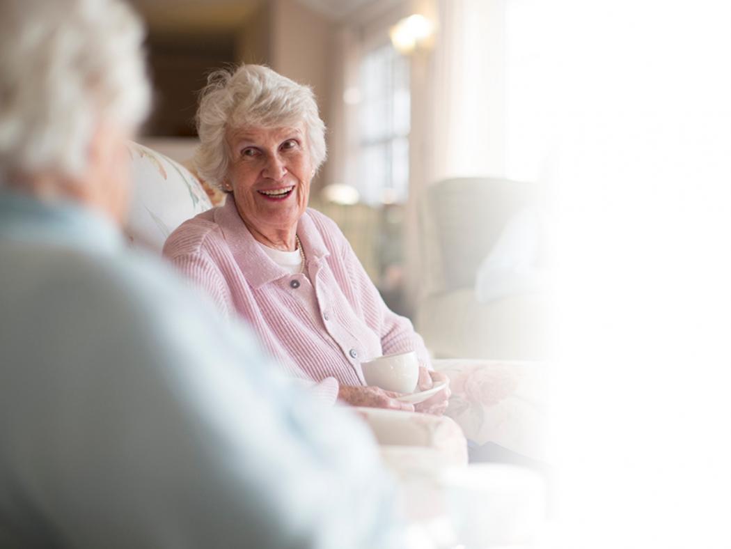 elderly lady sitting in a chair facing another elderly lady smiling