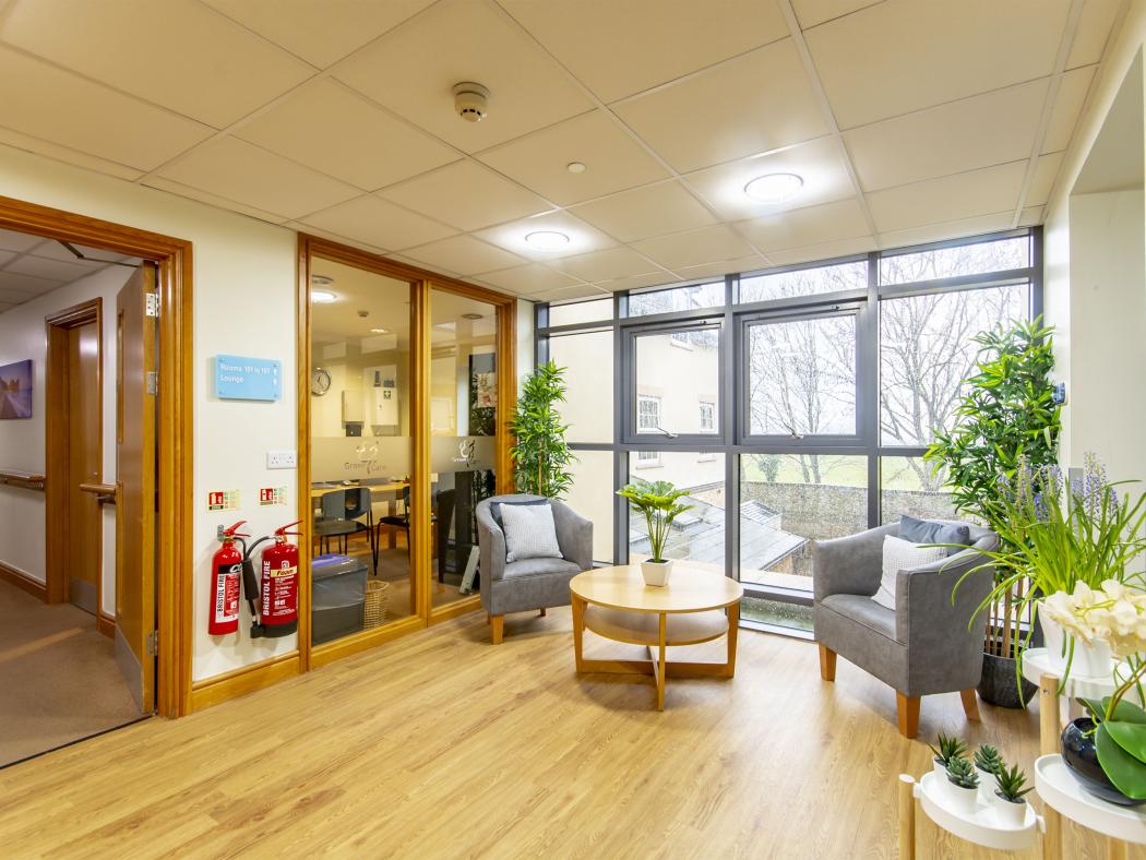 A bright lounge area with large windows, comfortable grey chairs, and decorative indoor plants, creating a welcoming atmosphere