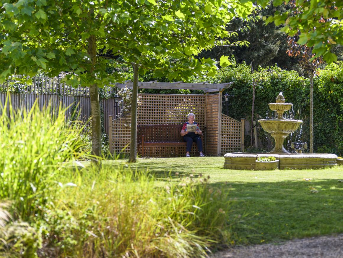 care home garden with water fountain and outdoors seating that resident is sitting on whilst reading 