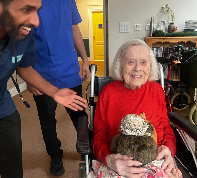 Smiling elderly woman in a red sweater holding a soft toy, seated in a wheelchair, surrounded by care staff